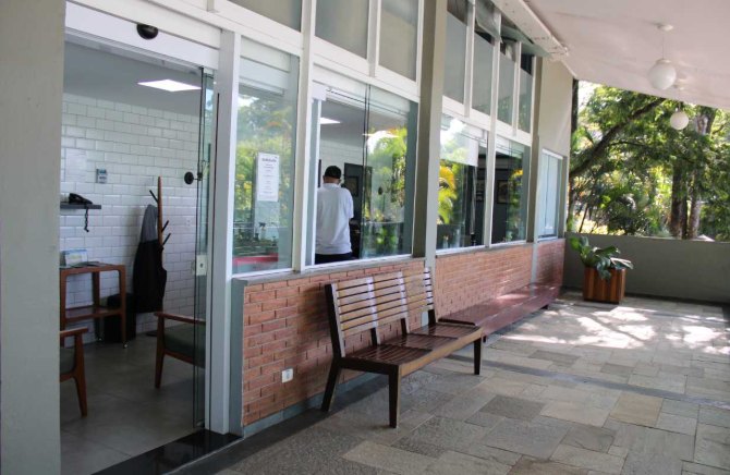 Entrada envidraçada da barbearia, com banco de madeira e piso de pedra, voltada para o jardim do clube.