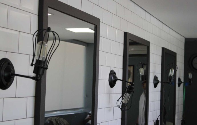 Espelhos de moldura preta e luminárias gaiola com lâmpada de filamento na parede de azulejo da barbearia.