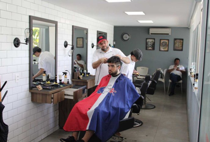Barbeiro cortando o cabelo de um cliente no salão da Barbearia ATC; ao fundo, outros clientes aguardam.